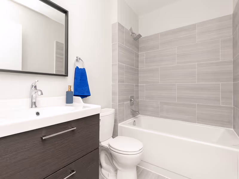 Modern bathroom with white bathtub, gray tiled shower wall, white toilet, dark wood vanity with sink, mirror, and blue towel.