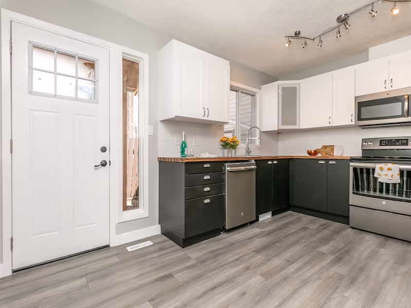 Modern kitchen with white upper cabinets, black lower cabinets, stainless steel dishwasher and stove, wooden countertop, and gray wood floor.