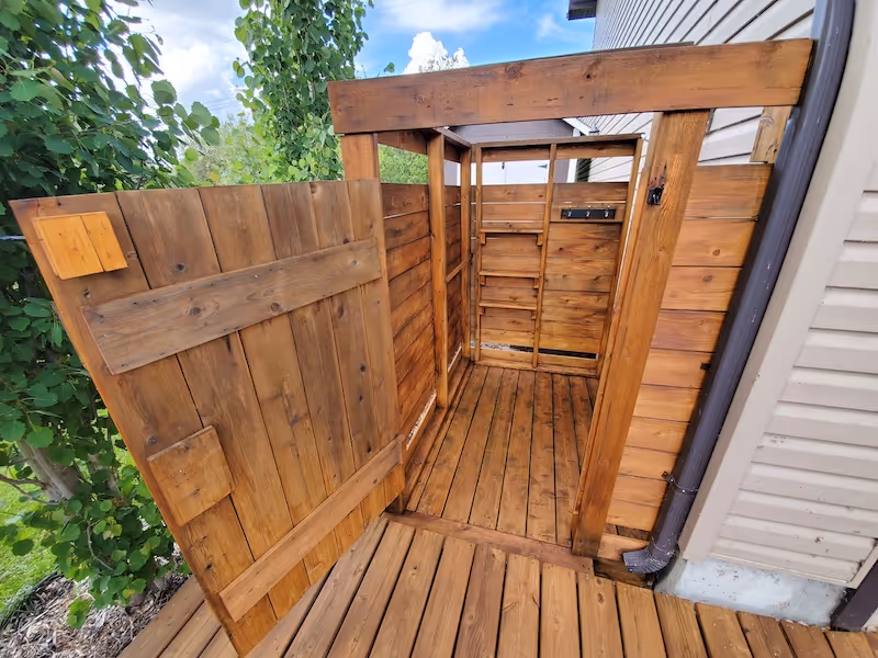 Open wooden outdoor shower enclosure next to a house with green trees and blue sky in the background.