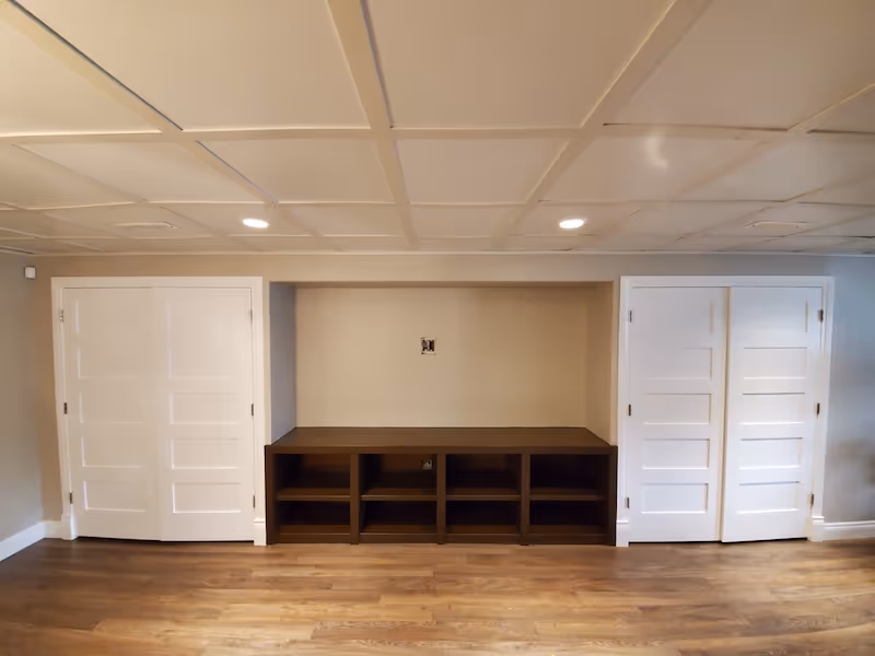Empty room with wooden floor, beige walls, ceiling lights, two white paneled doors on each side, and a dark wooden built-in shelf unit in the center.