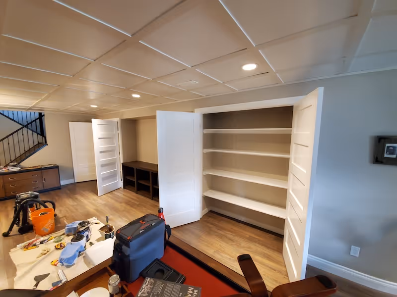 Room under renovation with tools, a vacuum, paint supplies on a table, an open empty white cabinet, and wooden flooring.