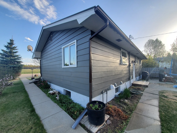 Exterior view of a single-story home with gray siding, white trim, landscaped garden beds, and a concrete walkway on a clear day.