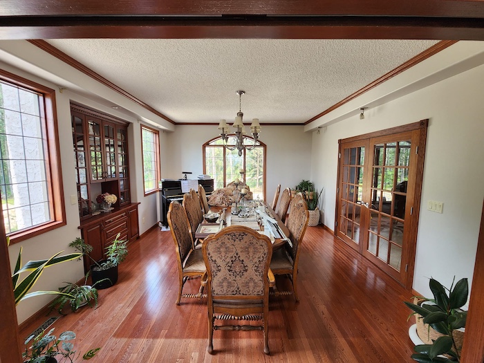 Elegant dining room with wooden floors, a long dining table, upholstered chairs, large windows, and decorative plants throughout the space.