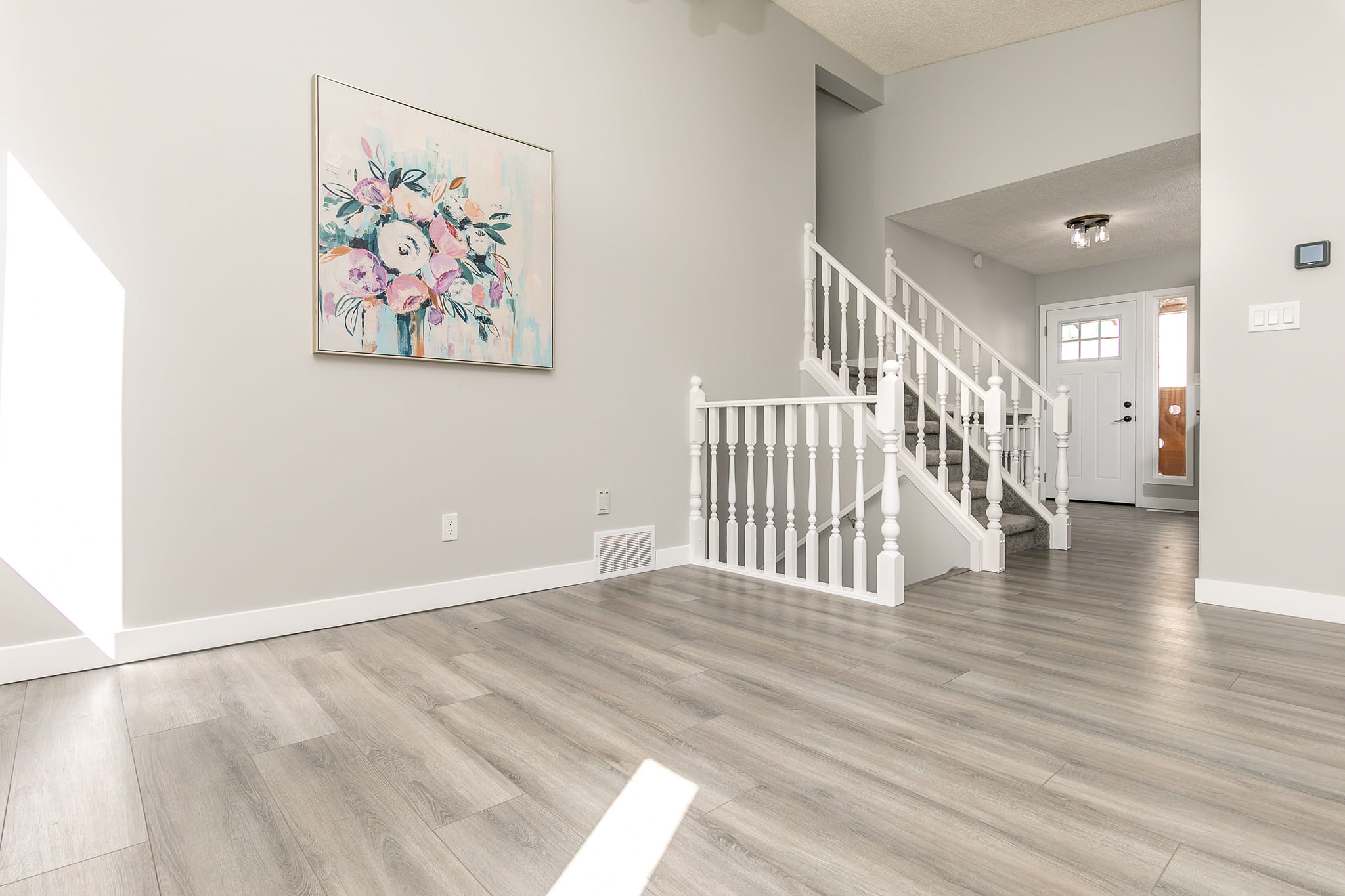 Bright open-concept room with light gray walls, wood-look flooring, a white railing staircase, and a floral artwork on the wall.