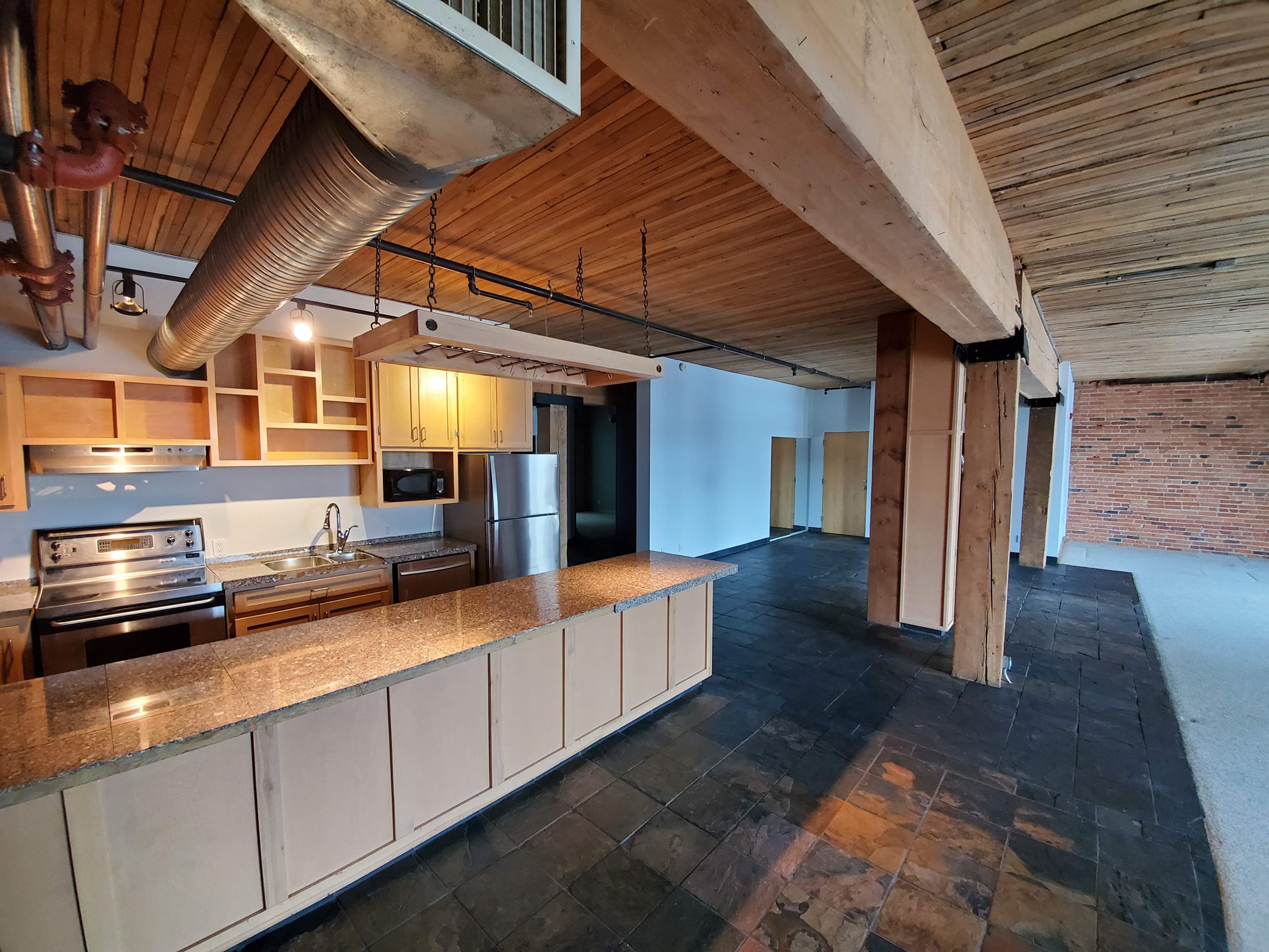 Rustic open-concept space with natural wood ceilings, exposed beams, slate flooring, and a kitchen with wood cabinetry and stainless steel appliances.