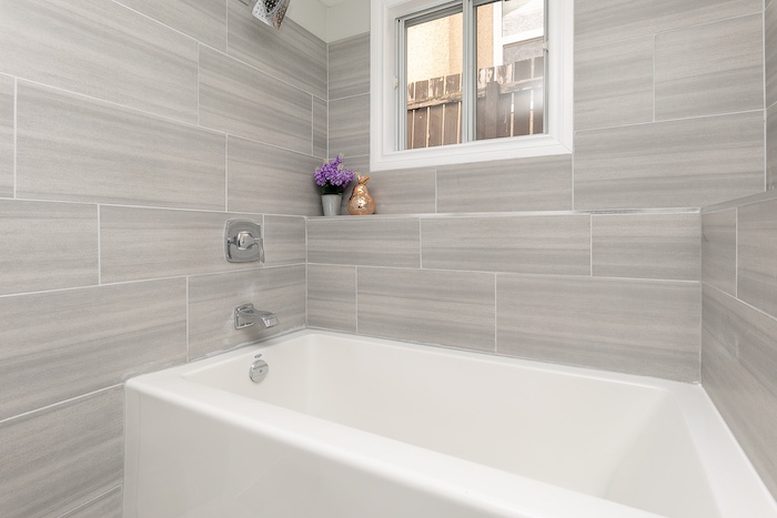 Bright bathroom with a white bathtub surrounded by light gray horizontal tile and a window above the tub.