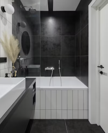 Modern bathroom with black tiled walls, a white bathtub with vertical paneling, a sleek vanity with vessel sink, and warm decorative accents.