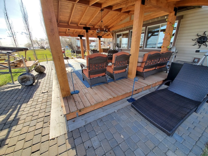 Covered outdoor patio with wooden posts and beams, cushioned seating, and stone paver flooring on a sunny day.