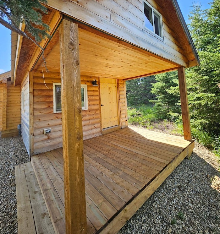 Small wooden porch with exposed beams and natural wood siding on a cabin-style building surrounded by trees.
