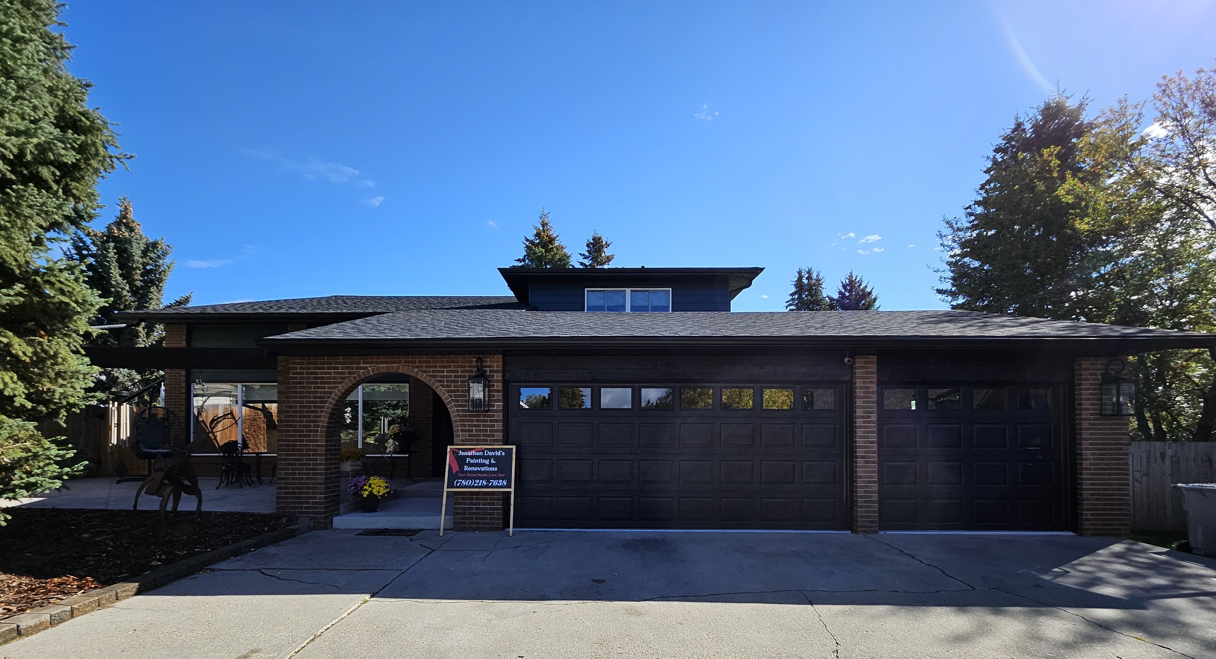 Front exterior view of a modern single-story home with dark siding, large garage doors, and a clean concrete driveway on a sunny day.