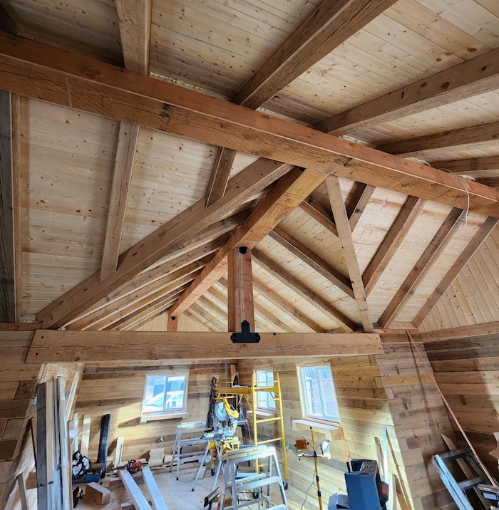 Interior of a timber-frame structure with exposed wooden beams, trusses, and construction tools throughout the workspace.