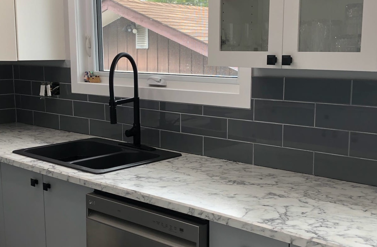 Modern kitchen counter with a marble-look surface, black faucet and sink, stainless steel dishwasher, and dark subway tile backsplash beneath a window.