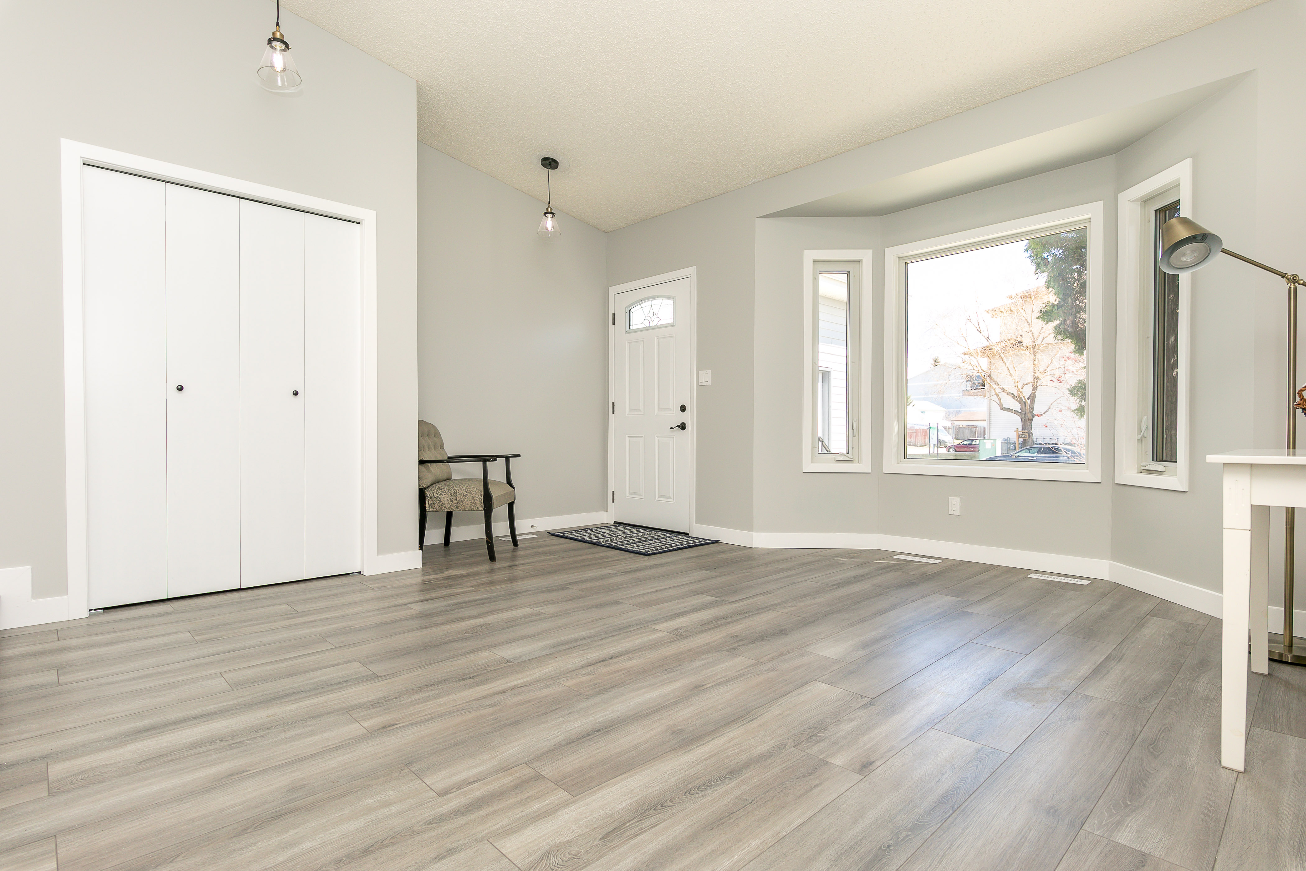 Bright, empty living room with light gray walls, wide plank flooring, a large bay window, and double white closet doors.