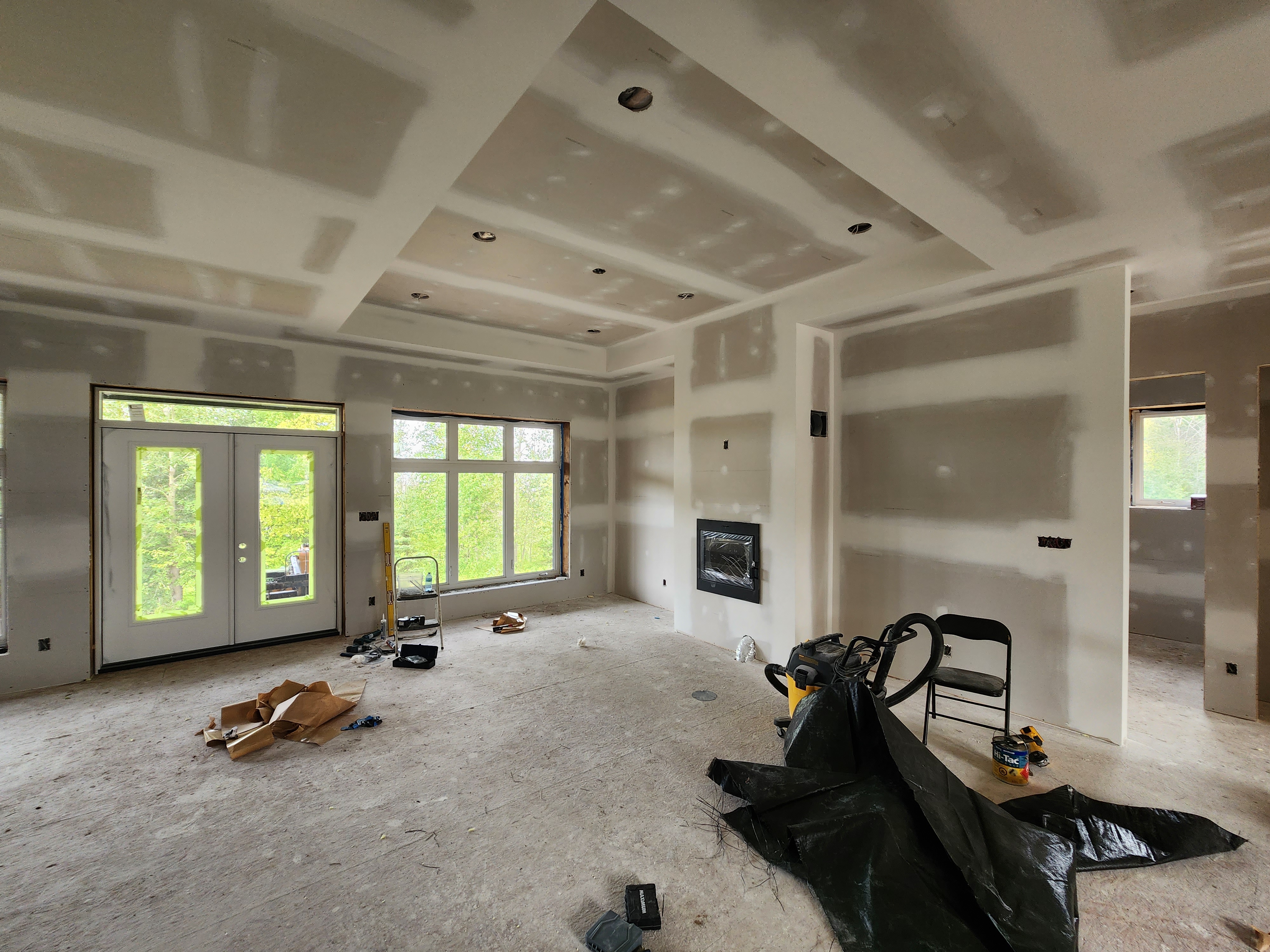Interior room under construction with unfinished drywall, exposed seams, tools on the floor, and large windows overlooking a green yard.
