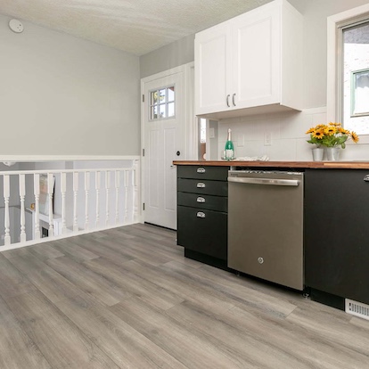 Modern kitchen with dark cabinetry, stainless steel dishwasher, light countertops, and gray wood-look flooring beside a white stair railing.