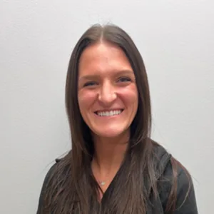 Woman with long brown hair smiling against a plain light gray background.