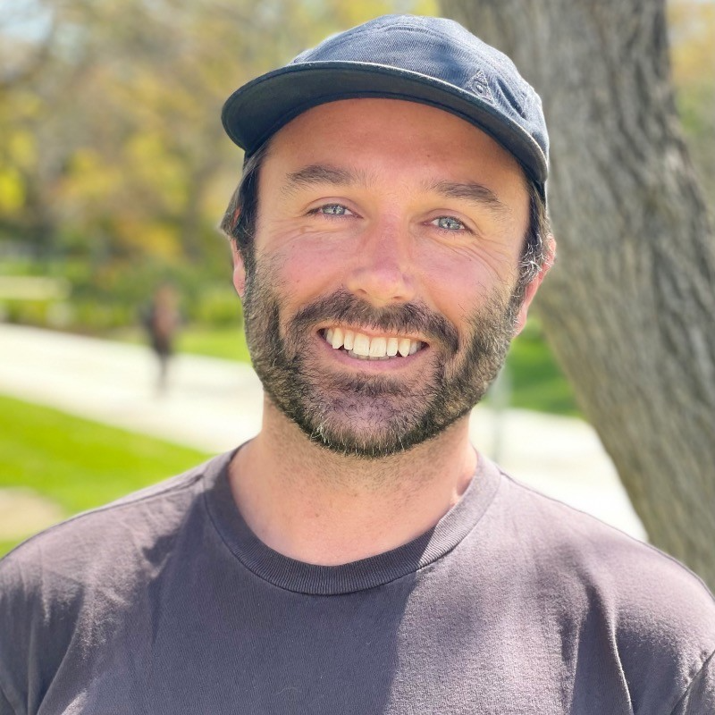 Smiling man with beard wearing a black baseball cap and dark shirt standing outdoors near a tree on a sunny day.