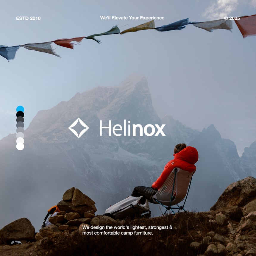 Person in a red jacket sitting on a Helinox camping chair on rocky terrain with misty mountains and prayer flags in the background.