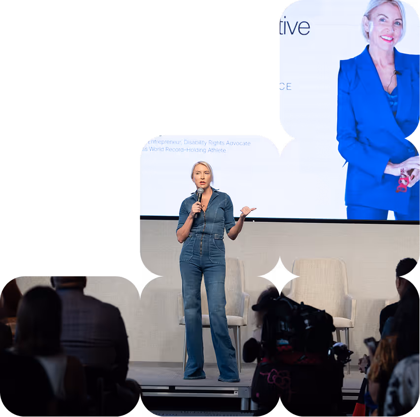 Woman in denim jumpsuit speaking on stage with microphone in front of seated audience and large screen displaying her image behind.