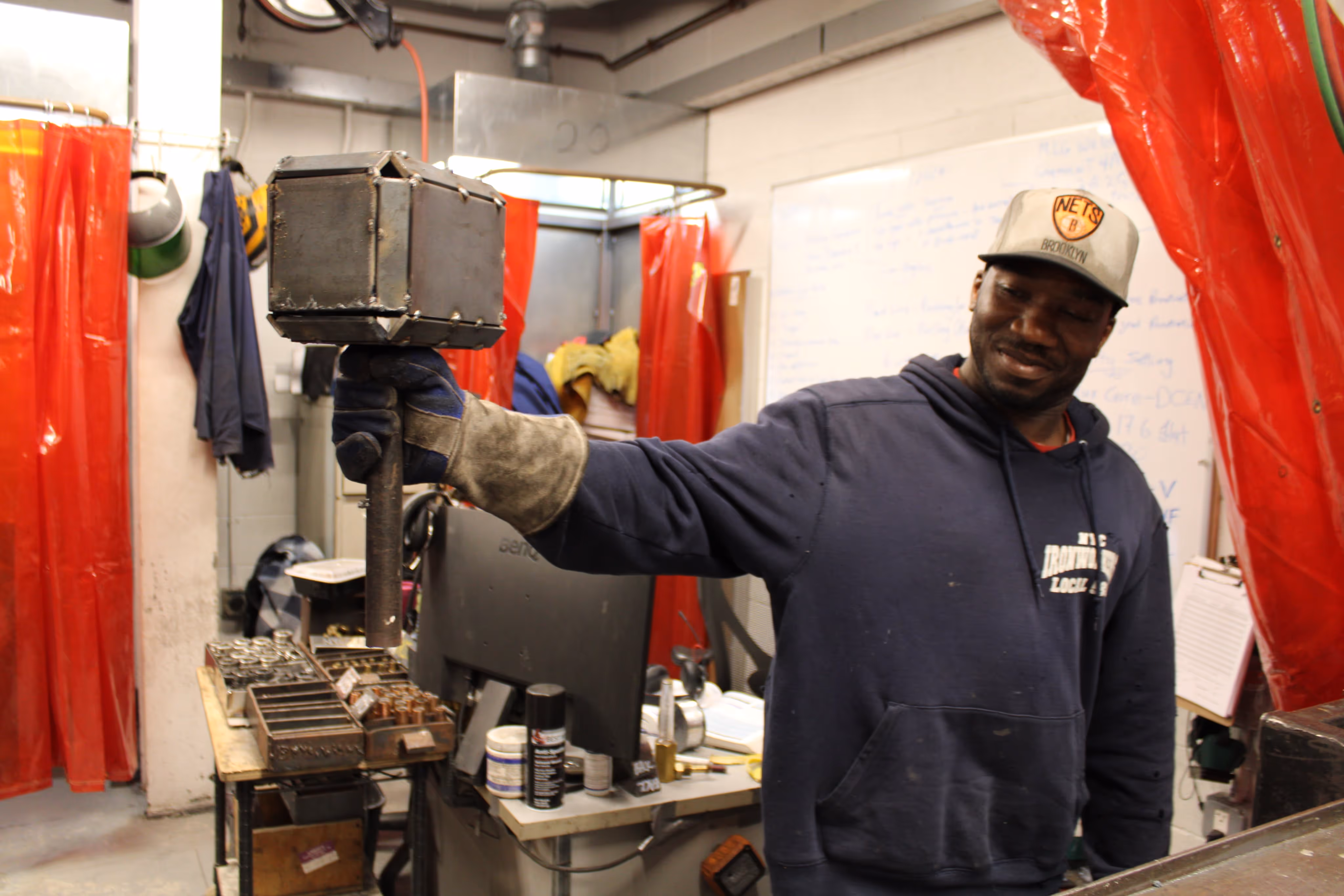 Worker in a workshop holding a large metal hammer and smiling, wearing gloves, a navy hoodie, and a Brooklyn Nets cap.