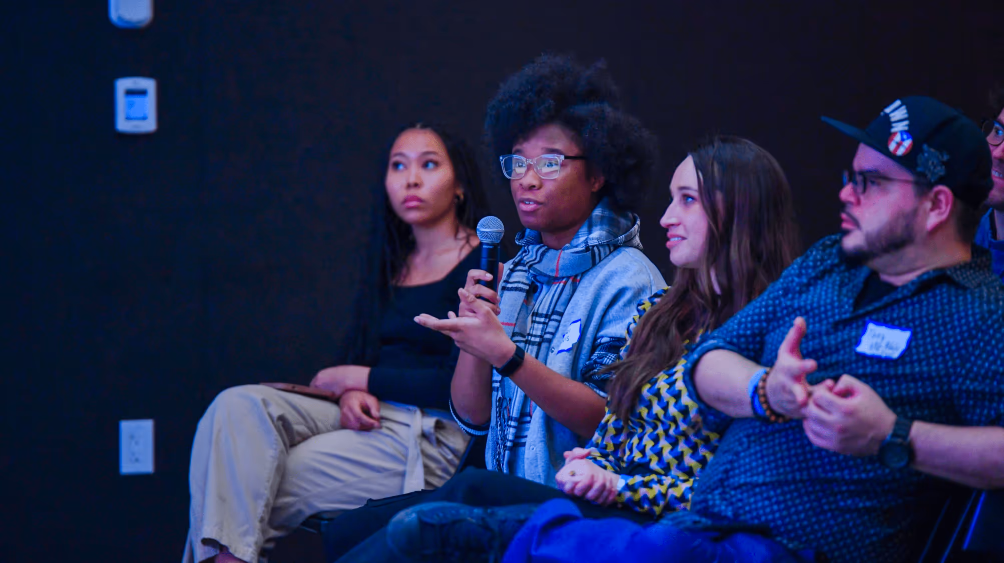 Group of diverse people seated, with one person holding a microphone and speaking during a discussion.