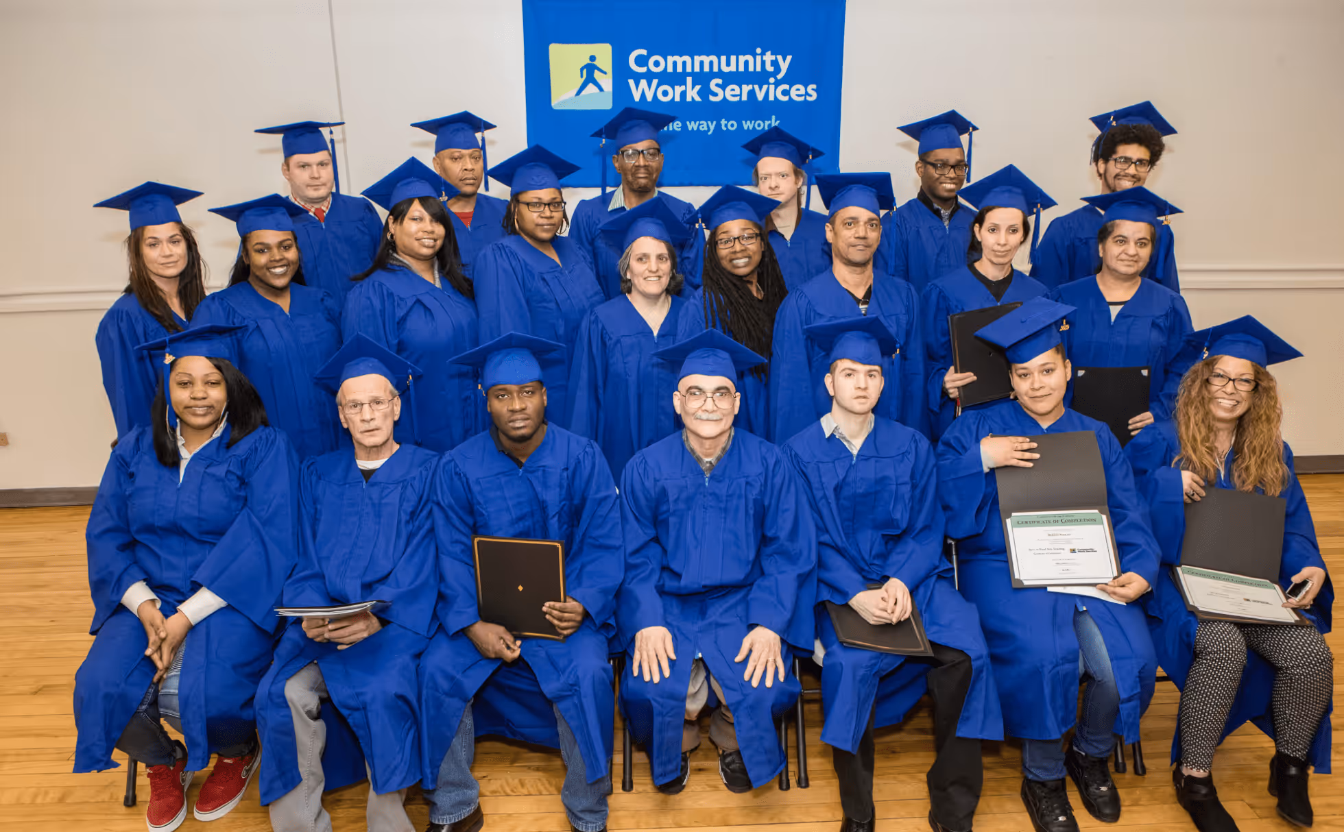 Group of diverse graduates in blue caps and gowns posing indoors with certificates in front of a Community Work Services banner.