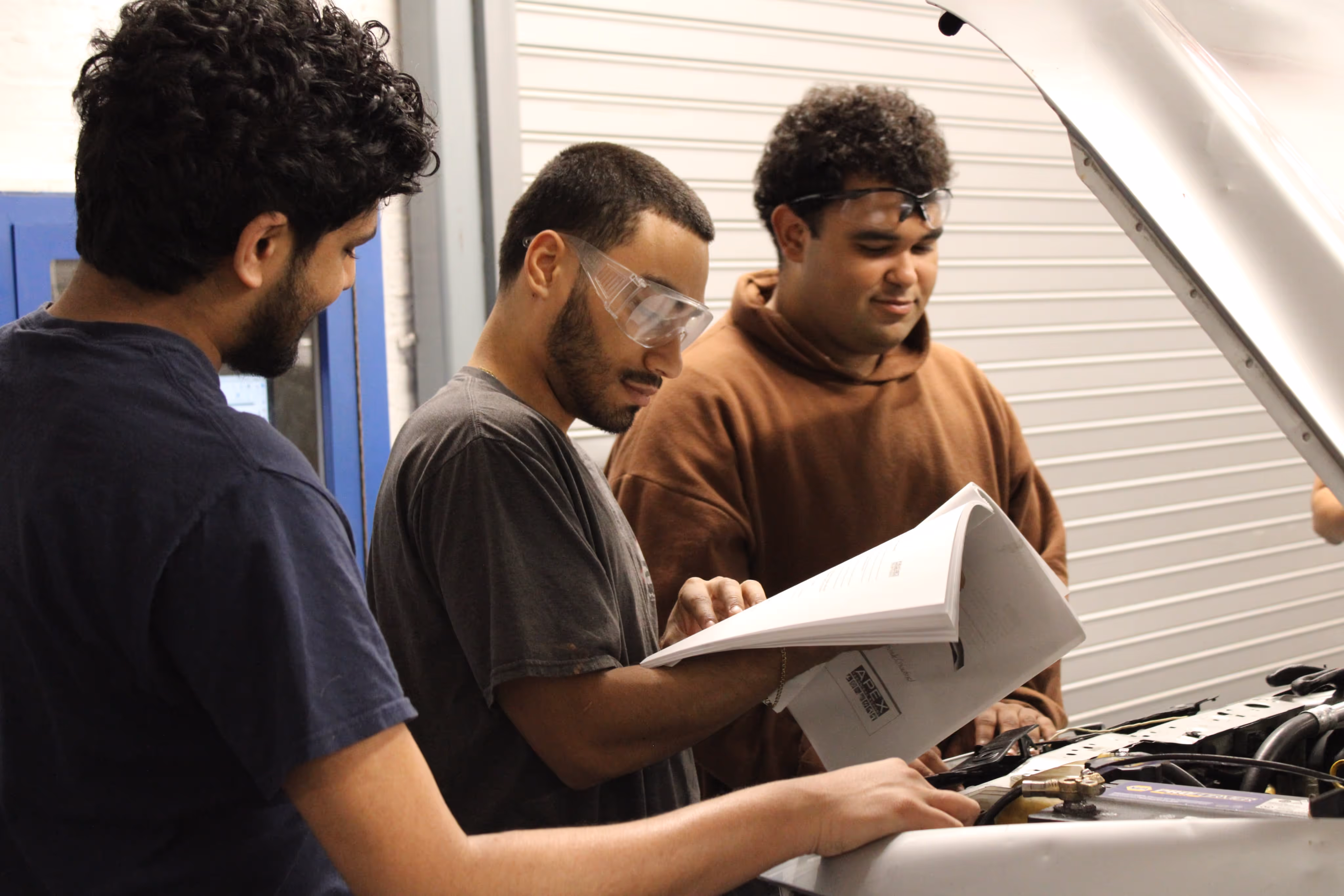 Three young men wearing safety glasses inspecting an open car hood and consulting a manual or guide.