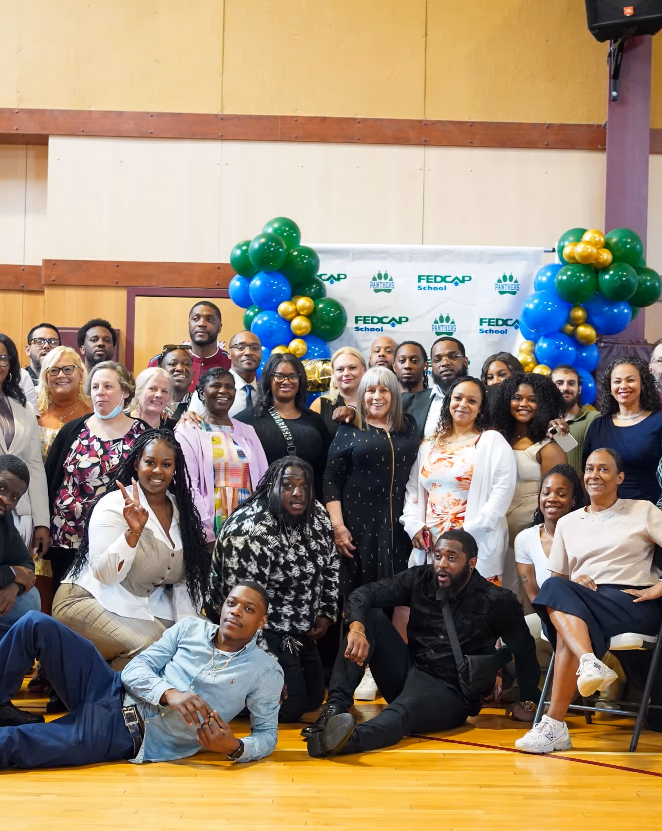 Group of diverse people posing indoors with blue, green, and gold balloon decorations and a backdrop featuring FEDCAP School and Panthers logos.