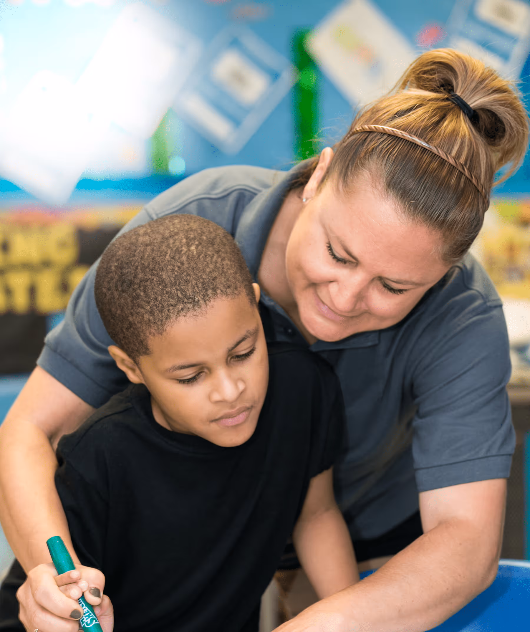 Woman with light skin and brown hair leaning over a boy with dark skin and short hair, both focused on an activity.