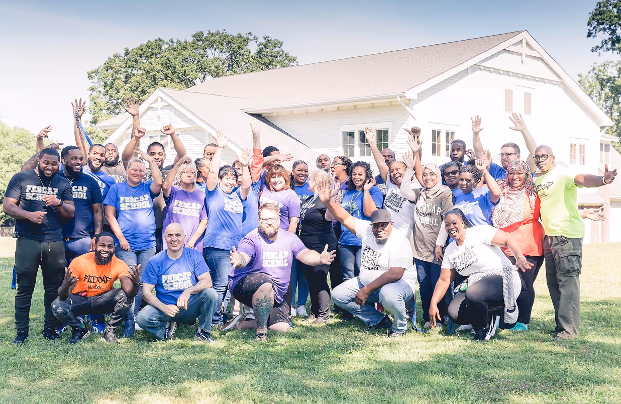 Group of diverse adults outdoors on grass in front of a white building, many wearing colorful shirts with raised hands and smiling.