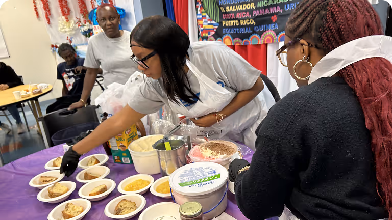 Two women serving food onto plates at a table covered with a purple tablecloth, with people and a colorful banner in the background.