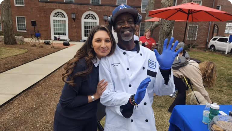 Smiling male chef wearing a white uniform and blue gloves stands next to a woman with long brown hair outdoors near a building with large windows.