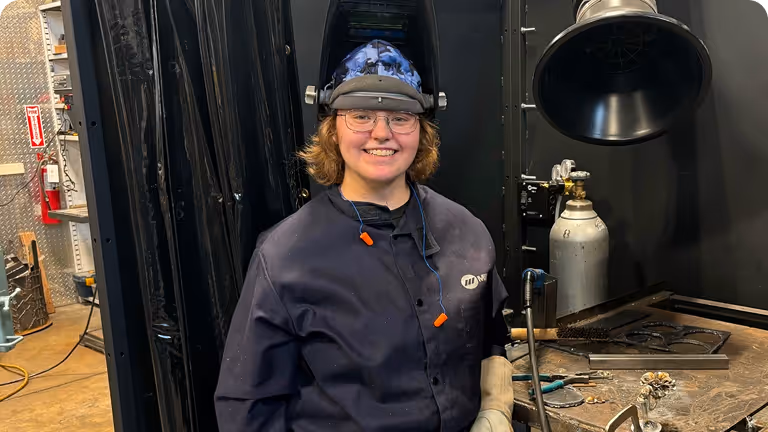 Smiling person in welding gear, including helmet and gloves, standing in a workshop with welding tools and equipment.