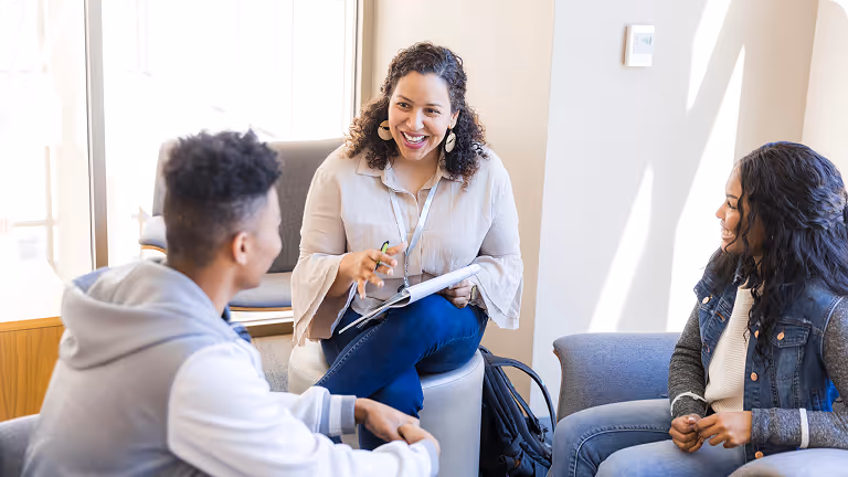 A smiling woman holding a notebook engages in conversation with two seated young adults in a bright room.
