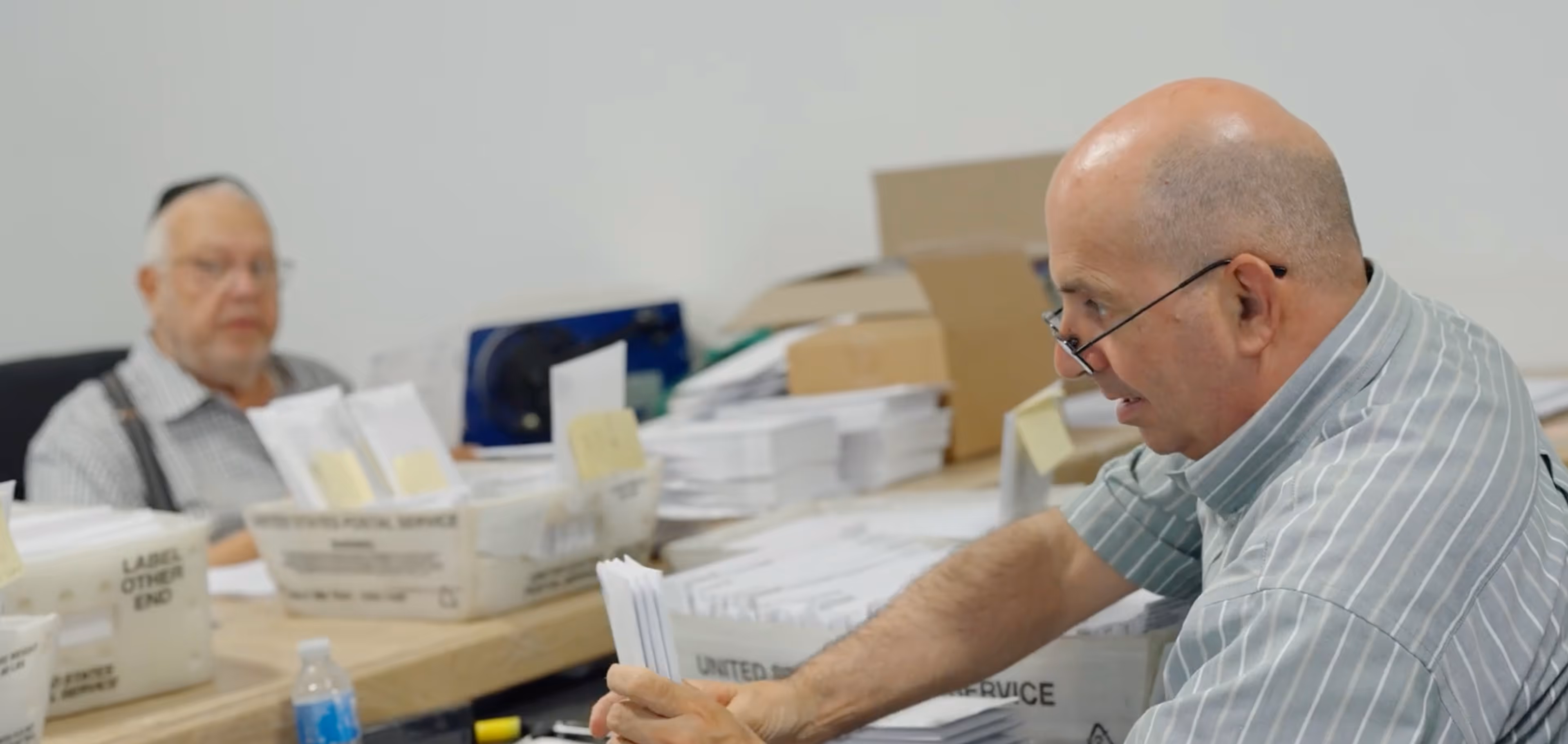 Technician wearing a cap and wireless earbuds inspecting and taking notes on a black Nissan Sentra in a busy auto repair shop.