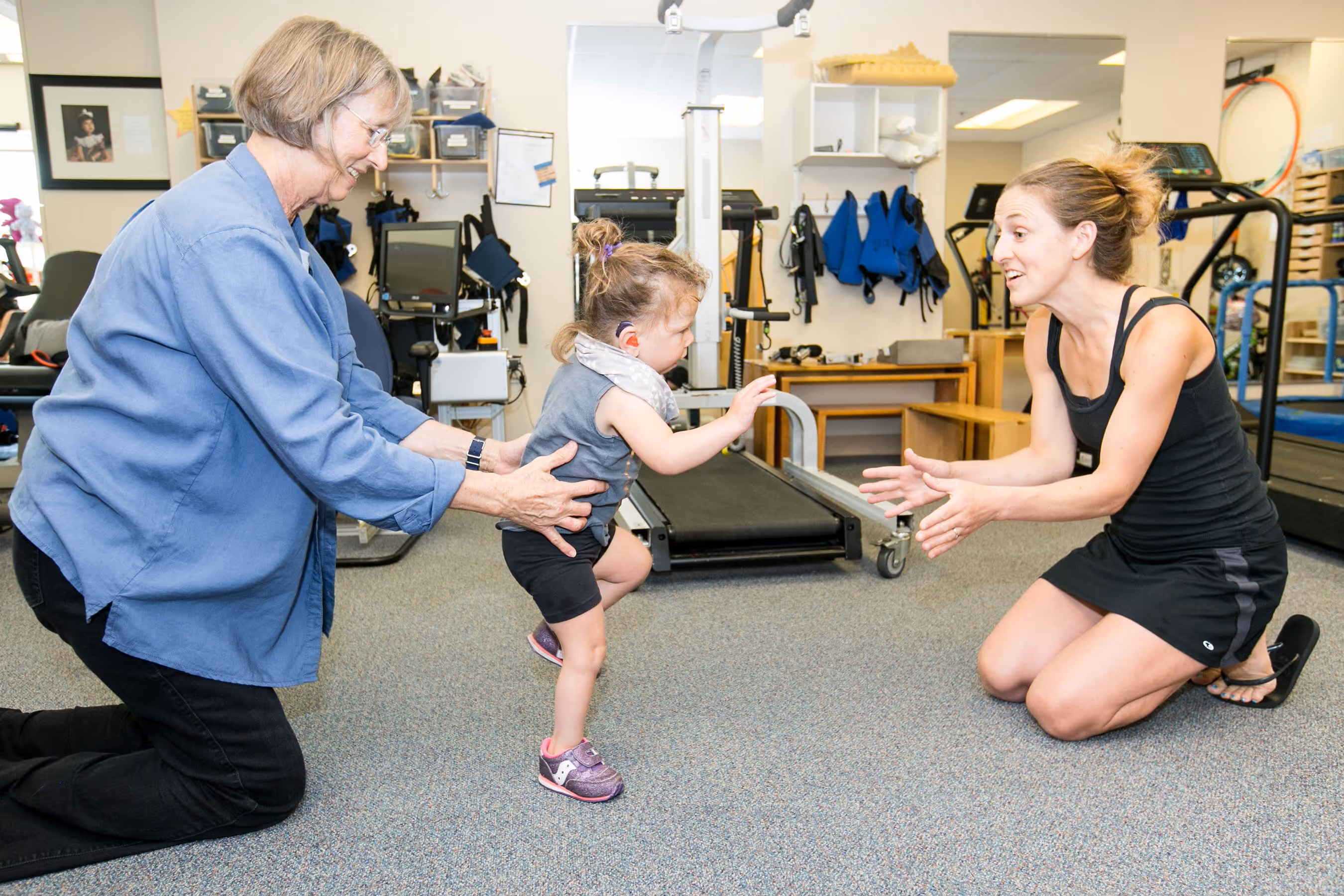 Child with hearing aid taking first steps toward a woman kneeling with open arms, while an older woman supports the child from behind in a therapy room.