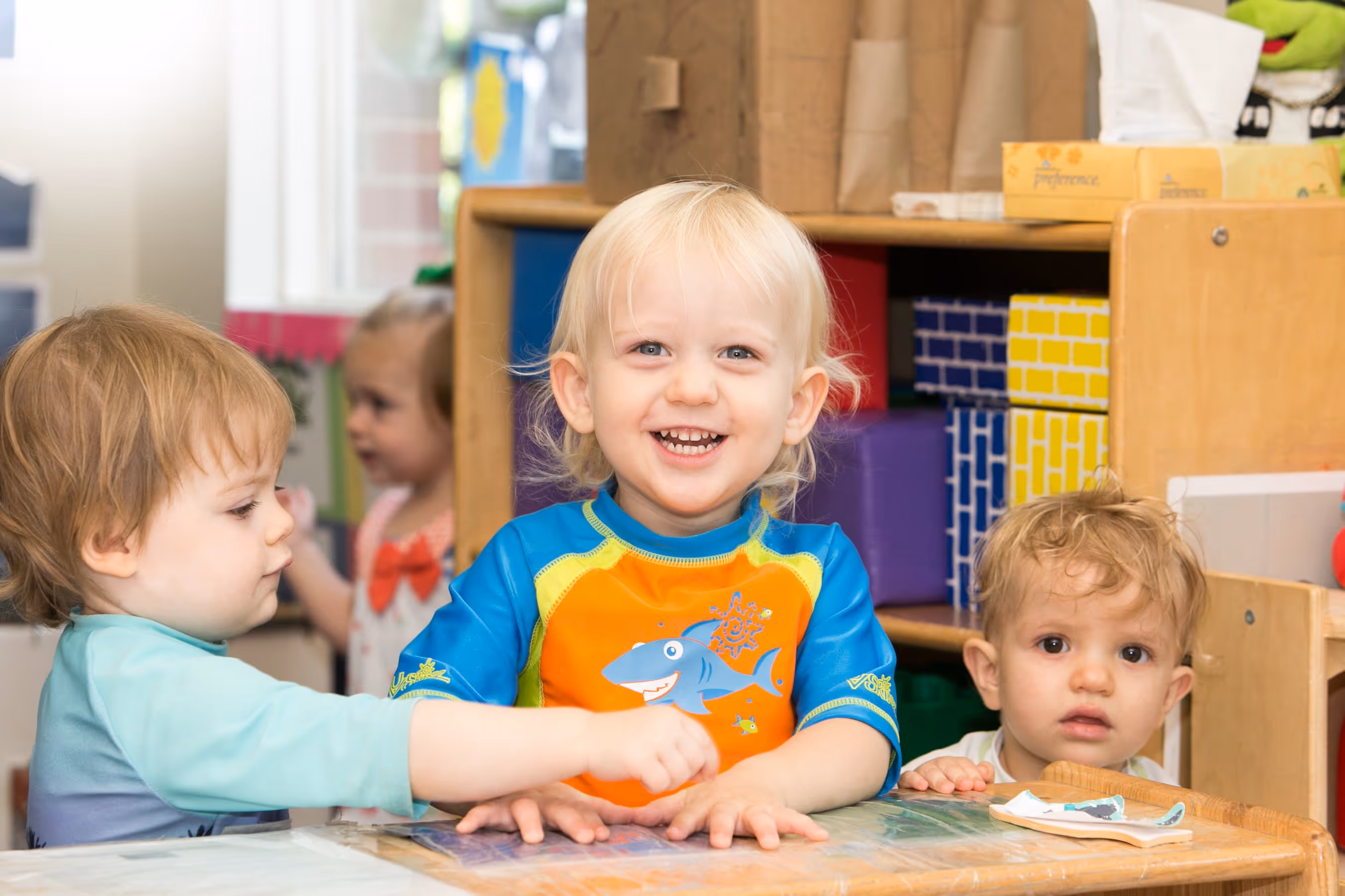 Three toddlers sitting and playing at a small table in a colorful classroom.