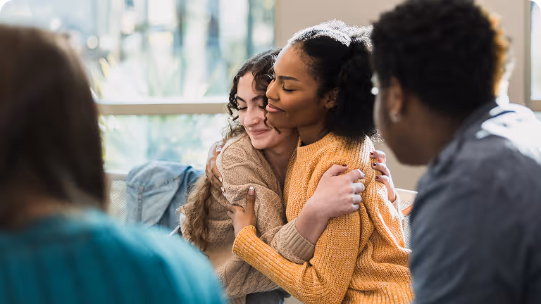 Two women warmly embracing each other in a group setting with two other people visible in the foreground.