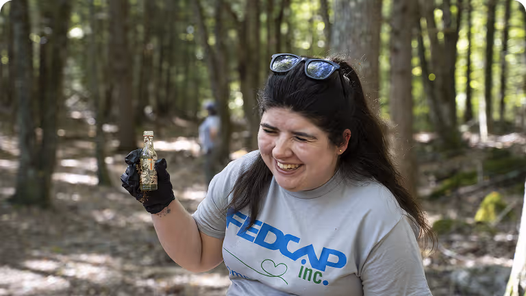 Smiling woman wearing a redcap inc. t-shirt and black gloves holding a small glass bottle in a wooded area.