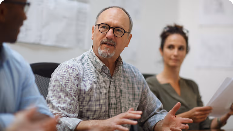 Middle-aged man with glasses speaking and gesturing his hands during a meeting with two colleagues.