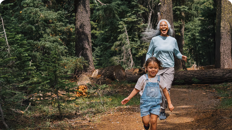 Smiling elderly woman and young girl running joyfully on a forest trail surrounded by tall trees.