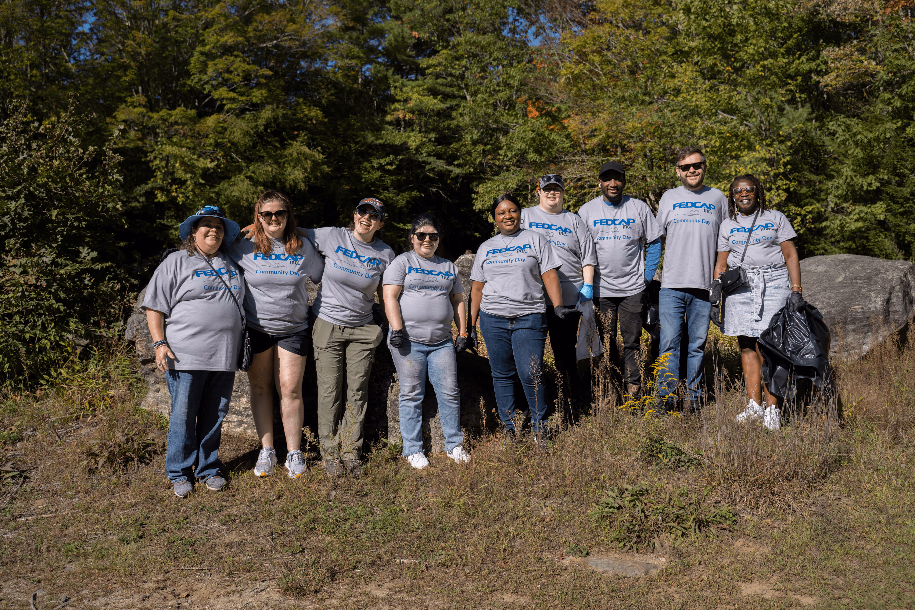 Group of nine people wearing matching gray FEDCAP Community Day shirts posing outdoors in front of trees and rocks.