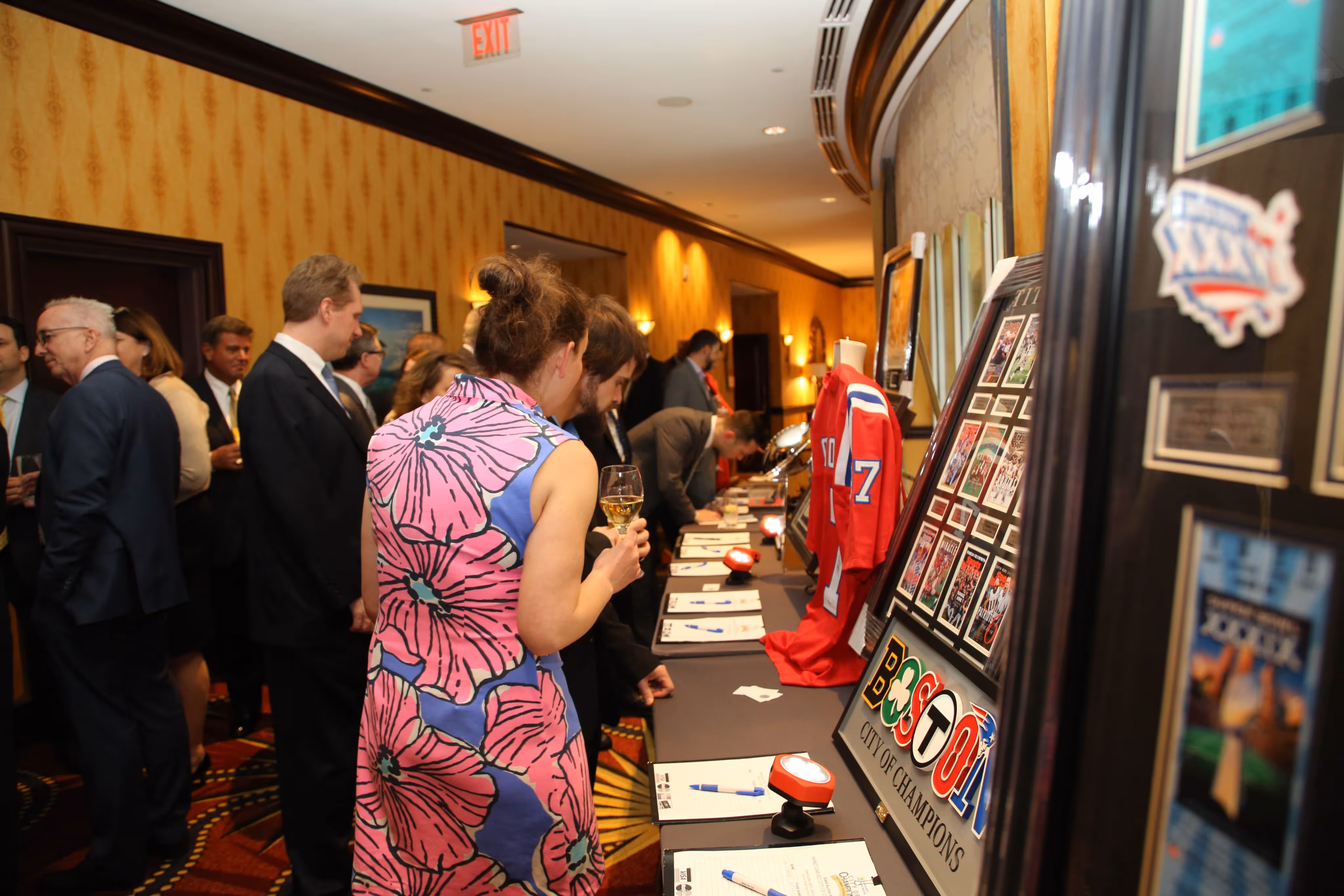 People in formal attire viewing and signing a silent auction table with sports memorabilia including a red jersey and framed photos in a carpeted hallway.