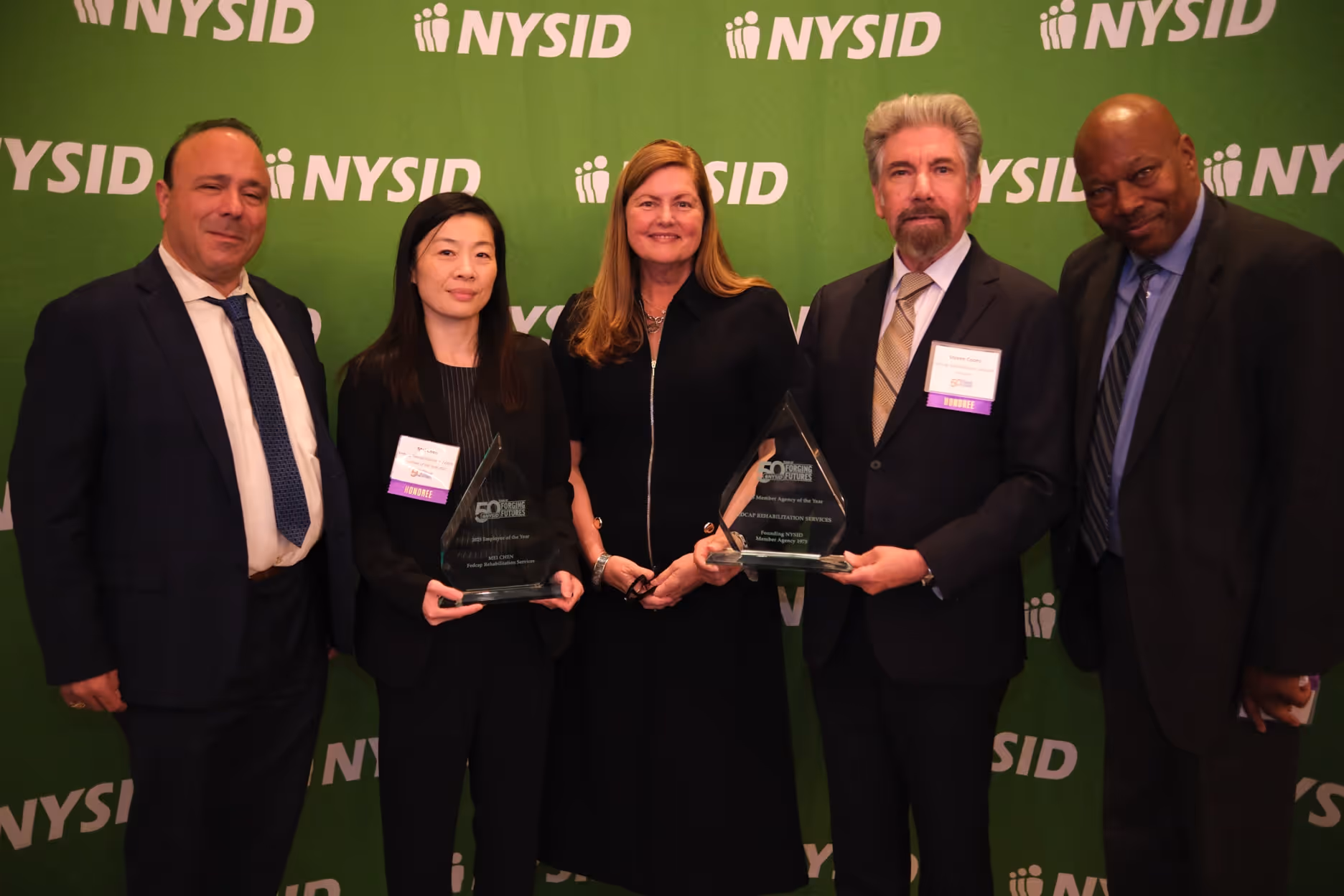 Five people in formal attire posing in front of a green NYSID backdrop, with two holding awards for Fedcap Rehabilitation Services.