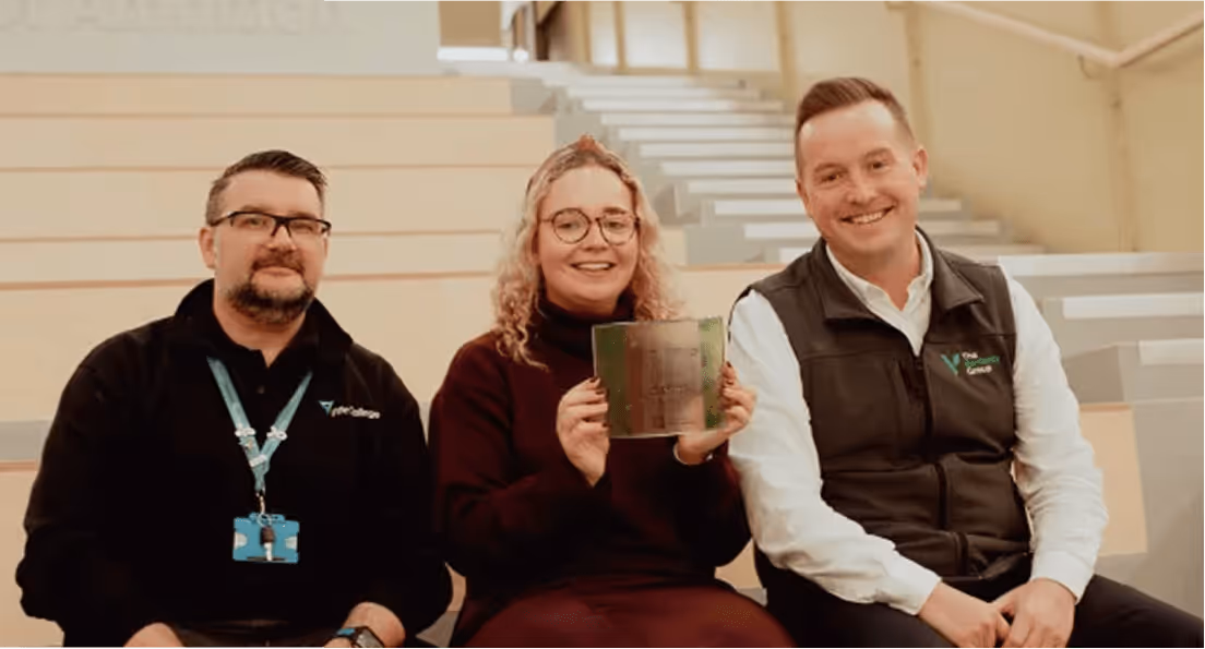 Three people sitting on stairs indoors, smiling; the woman in the center holds a glass award.
