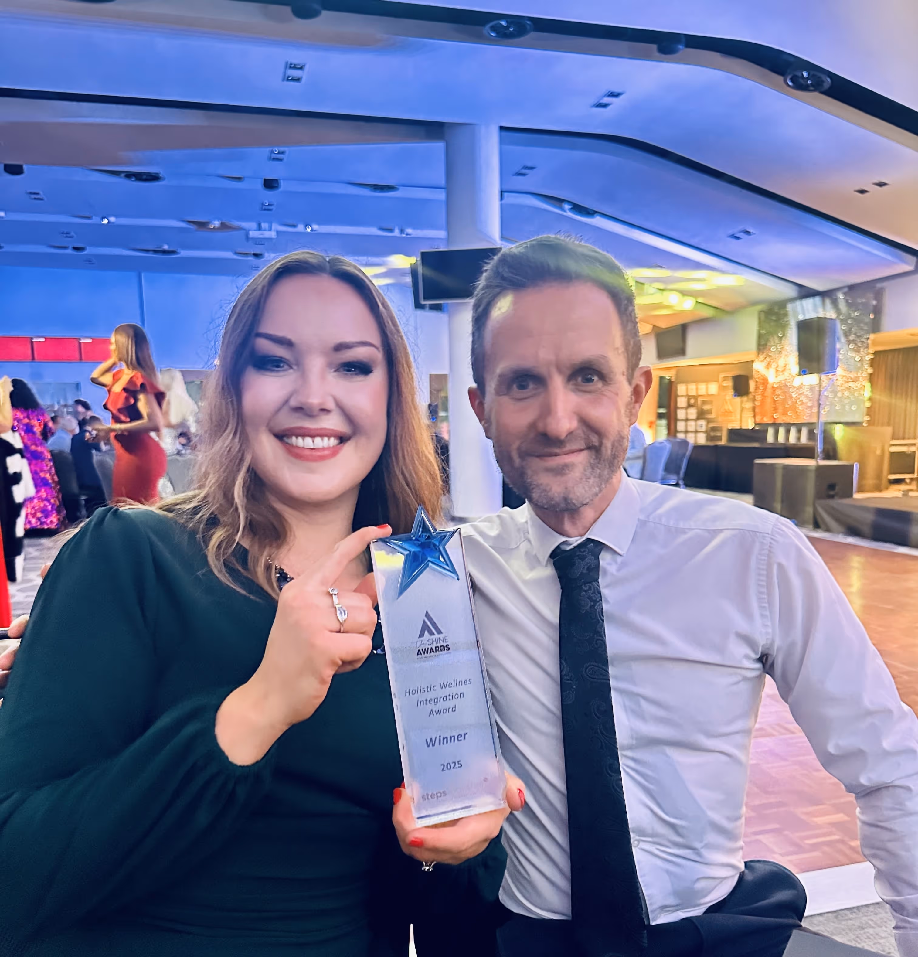 Smiling woman and man seated at an event holding a clear award with a blue star labeled 'Holistic Wellness Integration Award Winner 2025.'