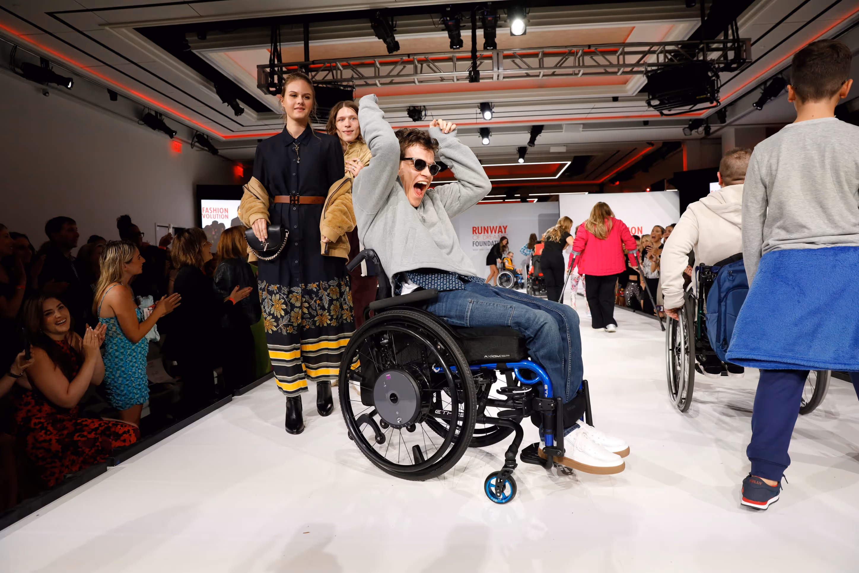 Person in a wheelchair cheering joyfully on a fashion runway with other models and spectators applauding.