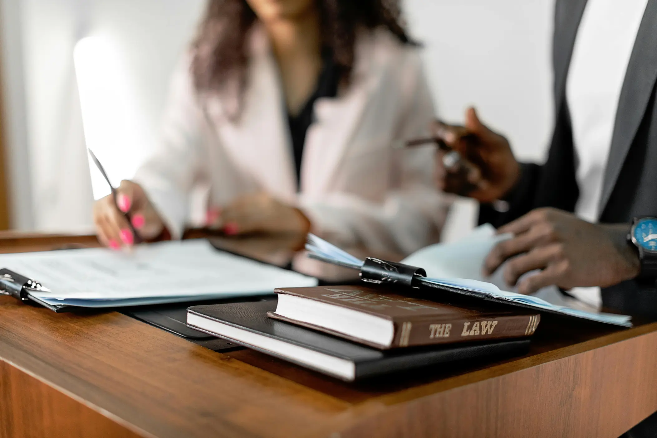 Une pile de documents posés sur un bureau avec un livre de droit et en arrière plan flouté une jeune femme portant une veste rose feuilletant un contrat. 