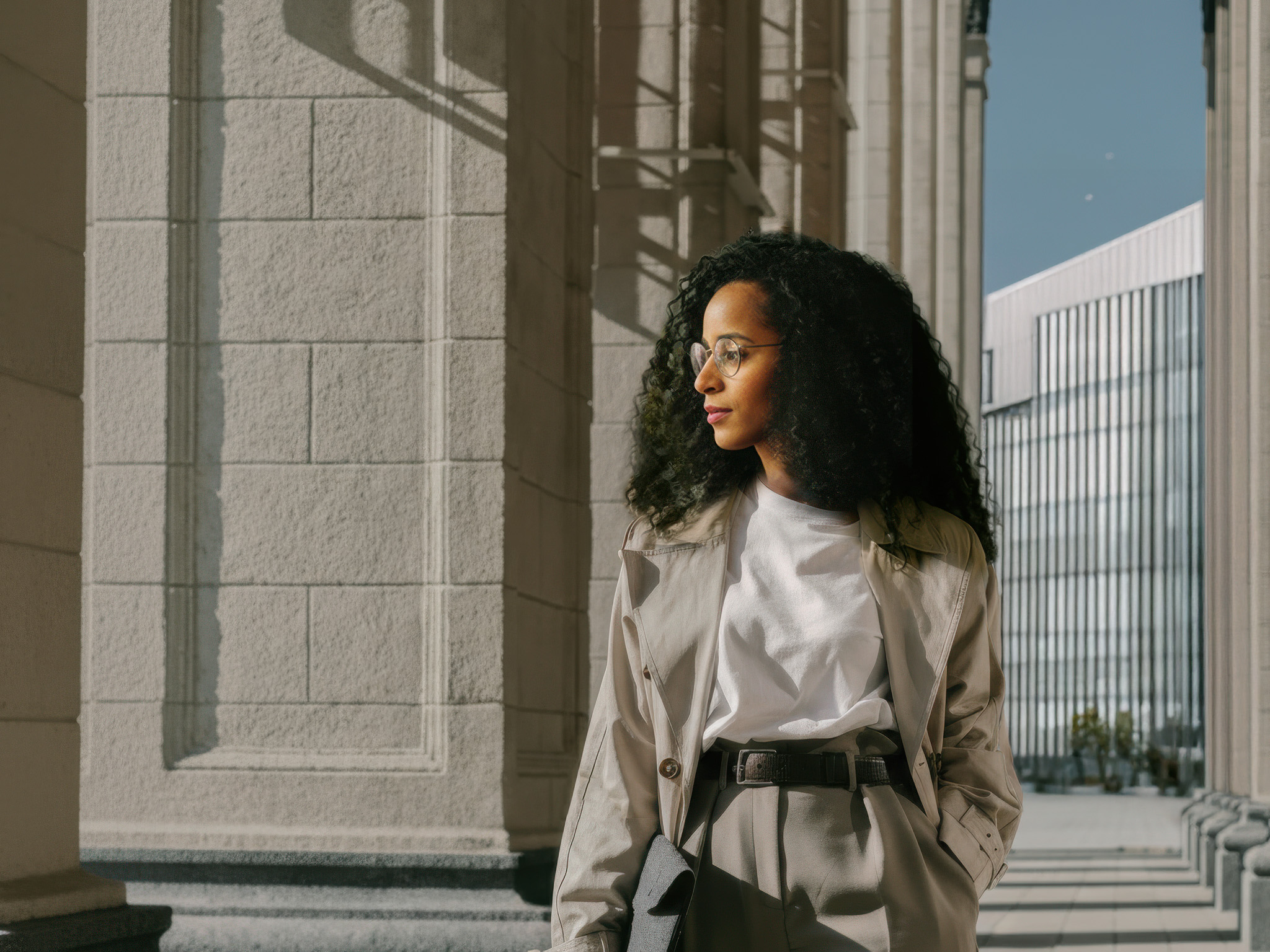 Jeune femme portant un trench-coat beige et des lunettes, debout devant un bâtiment moderne en pierre sous un ciel bleu.
