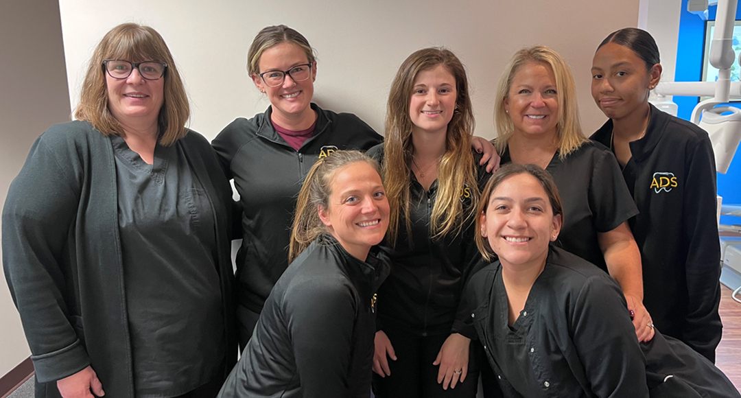 Group of seven smiling women posing indoors, some wearing black ADS-branded uniforms.